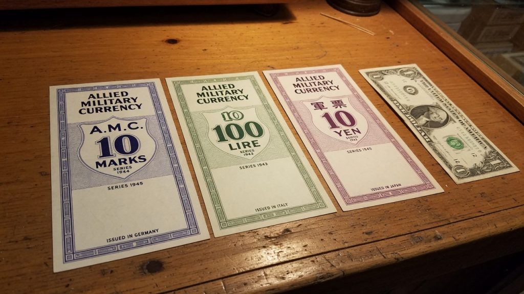 Three Allied Military Currency notes — marks, lire, and yen — beside a US dollar