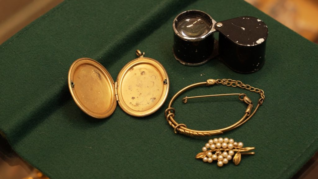 An open locket, a gold-colored bracelet, a pearl brooch, and a loupe on a green coin-shop pad