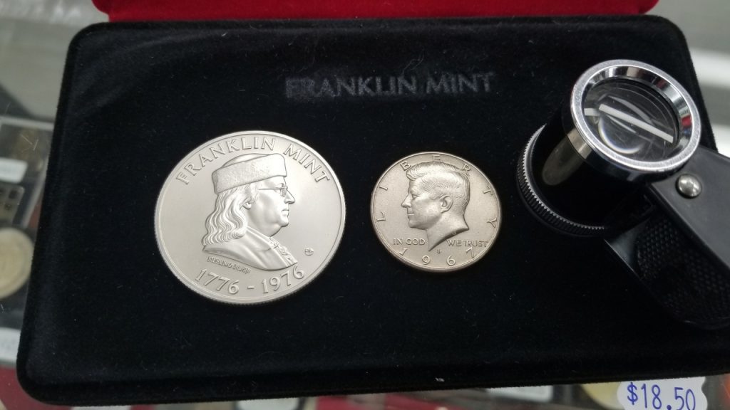 A Franklin Mint sterling medal in a case next to a Kennedy half dollar and a loupe