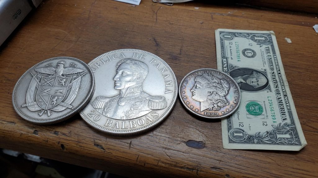 A large Panama 20 balboas silver coin next to a Morgan dollar and a one-dollar bill
