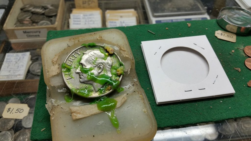 A dime with green PVC residue in a plastic flip beside a cardboard 2x2