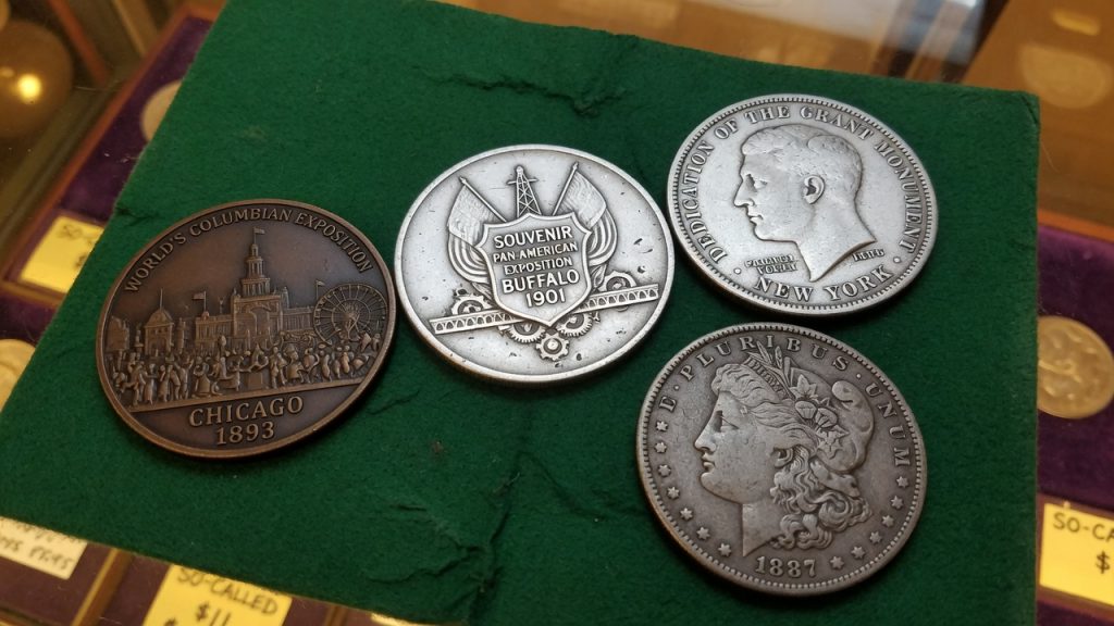 Dollar-sized exposition medals next to a Morgan silver dollar on a green pad