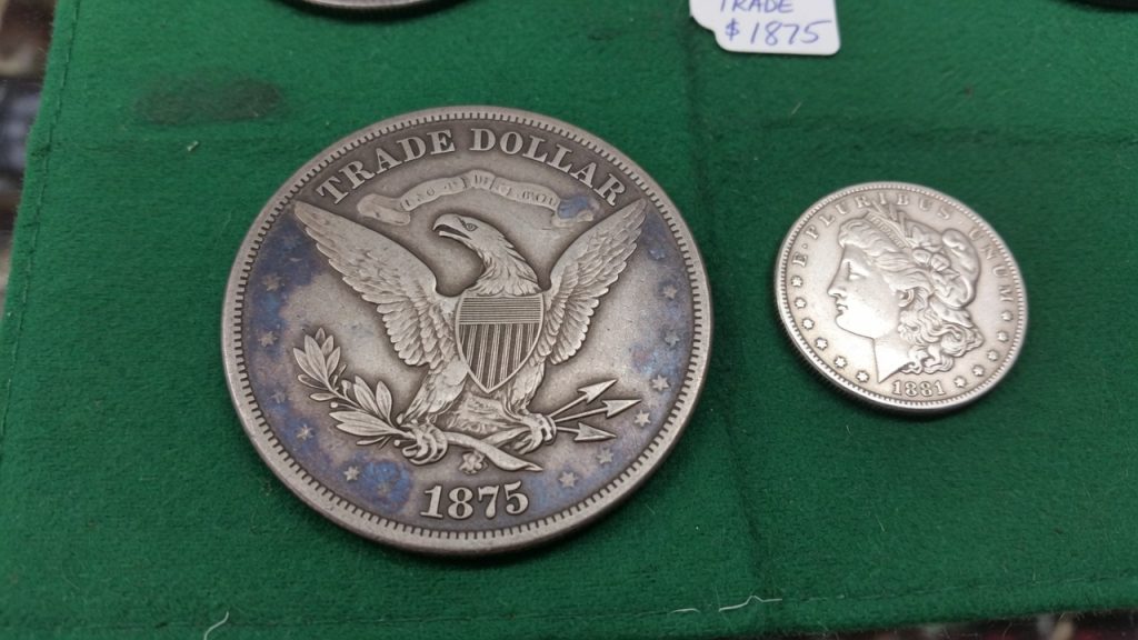 An 1875 Trade dollar reverse beside a smaller Morgan dollar on green felt
